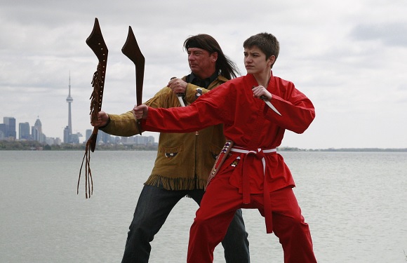 Two individuals practicing martial arts with traditional weapons by a lakeside with a city skyline in the background.