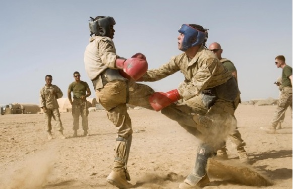 Two military personnel are sparring with boxing gloves in a desert setting, with other soldiers watching in the background.