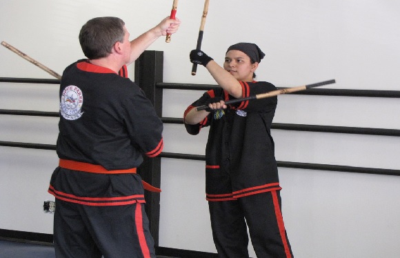 Two individuals in martial arts uniforms practicing with sticks in a gym-like setting.