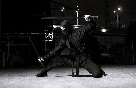 A person in dark clothing performing a martial arts stance with a sword, in a low-lit outdoor setting at night.