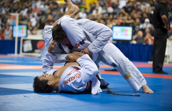 Two people in white gis are practicing Brazilian Jiu-Jitsu on a blue mat, with spectators watching.