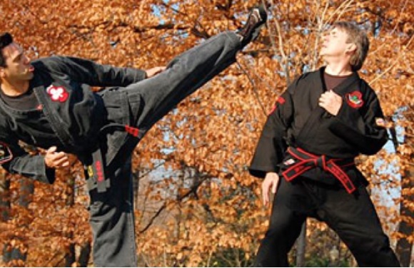 Two people in black uniforms and red belts are practicing martial arts outdoors, amidst autumn leaves.