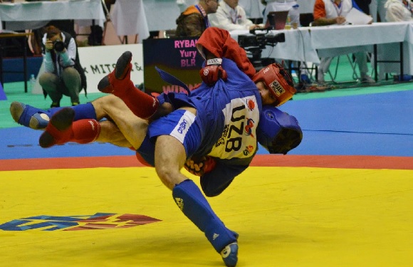 Two martial artists are fighting on a yellow mat, one doing a high kick, as spectators watch.