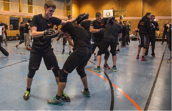 People are practicing martial arts in a gym, with two individuals sparring in close combat in the foreground.