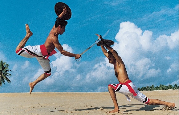 Two men are on a beach practicing martial arts; one jumps with a hat, the other crouches with a stick, both in striped pants.