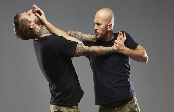 Two men practicing self-defense or martial arts, one performing an arm lock on the other against a grey background.