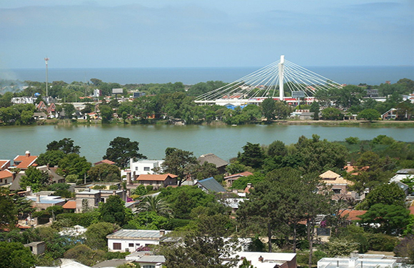 Aerial view of a town with a river, a white cable-stayed bridge in the background, and lush greenery surrounding the area.