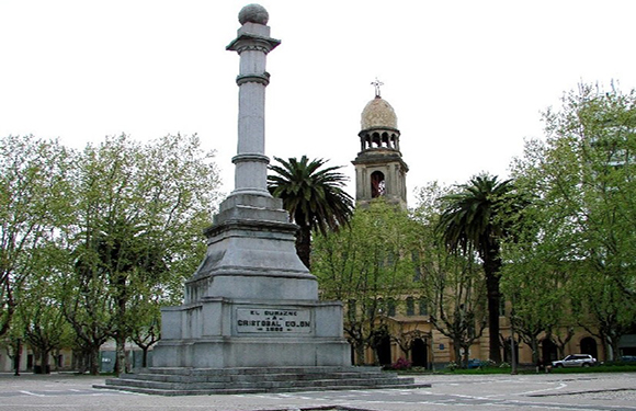 A monument consisting of a tall column on a square base, surrounded by trees with a church tower visible in the background.