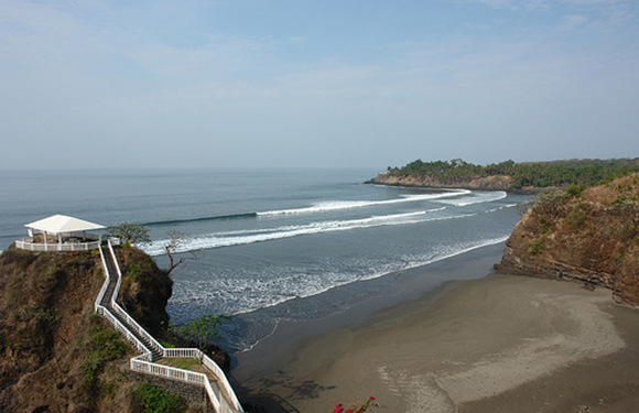 Aerial view of a serene coastal landscape featuring a beach, calm sea, pathway to shore, and a small pavilion.