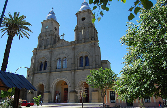 A photograph of a large church with two bell towers and a cross on top, situated under a clear blue sky with palm trees in the foreground.