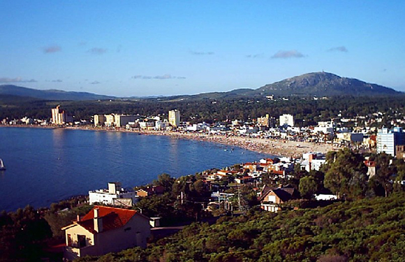 Aerial view of a coastal city with buildings along the shore, a beachfront, and a hill in the background under a blue sky.