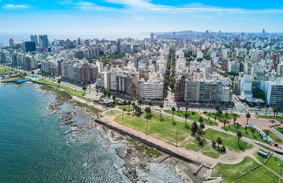 Aerial view of a coastal city with dense building clusters, green park areas, and a shoreline with waves hitting the rocks.