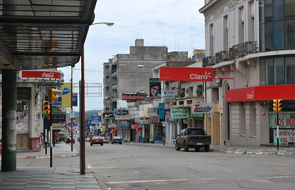 A street view with various storefronts, signage, and a few cars parked and driving on the road, under an overcast sky.