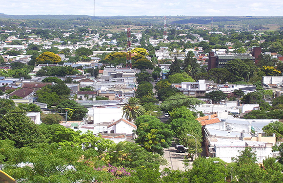 Aerial view of a small city with dense building clusters, green trees scattered throughout, and a flat horizon in the background.