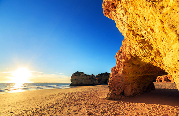 A scenic beach during sunset with a large rock formation on the left and smaller formations in the distance under a clear sky.