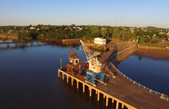 Aerial view of a river with a dock and a large industrial crane on it, with a boat moored alongside and a town in the background under a clear sky.