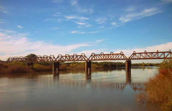 An image of a long, metal truss bridge spanning over a calm body of water with greenery on the banks under a blue sky with light clouds.