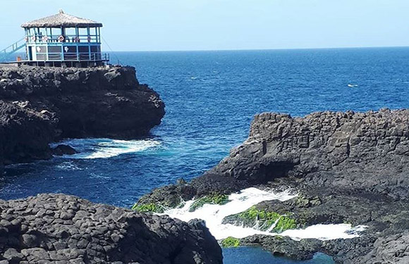 A coastal landscape with a gazebo structure on a cliff overlooking the sea, with rocky formations and waves crashing below.