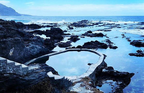 A natural rock pool by the sea with a curved white border, surrounded by dark volcanic rocks against a backdrop of the ocean and a cloudy sky.