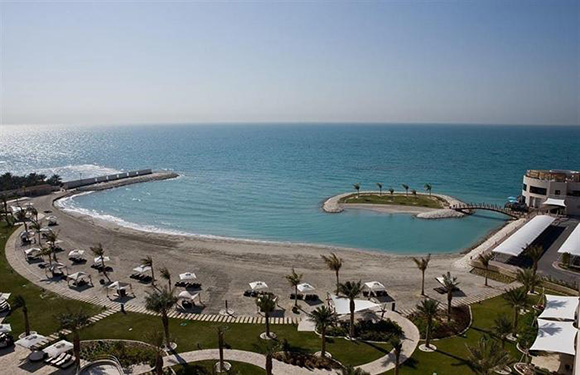 Aerial view of a coastal area with a curved beach, calm blue sea, palm trees, and a grassy area with umbrellas and sun loungers.