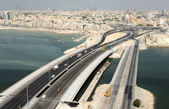 Aerial view of a multi-lane highway bridge over water with city skyline in the background.