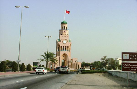 A photograph of a clock tower with a flag at the top, situated by a road with cars and street signs, with trees and lampposts in the background.