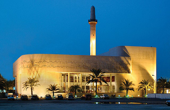 An image of a modern mosque with a tall minaret at dusk, illuminated by exterior lights, with palm trees in the foreground.