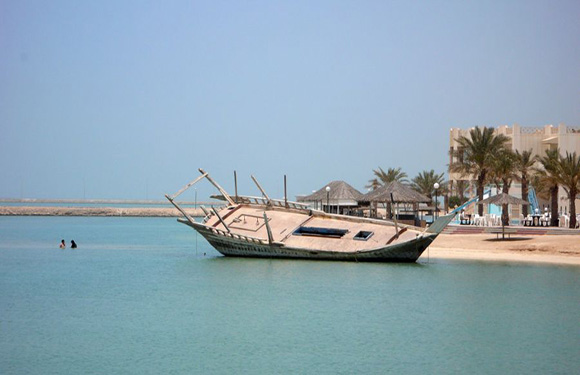 A traditional dhow is beached on a shore with calm waters, clear sky, nearby buildings, palm trees, and two swimmers.