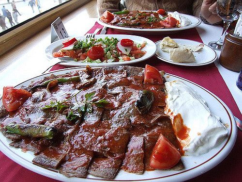 A plate of Turkish doner kebab with slices of tomato and a dollop of yogurt on the side, accompanied by other dishes in the background on a table.