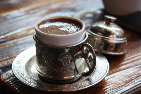 A traditional cup of coffee in a metal cup and saucer with intricate designs, placed on a wooden surface.