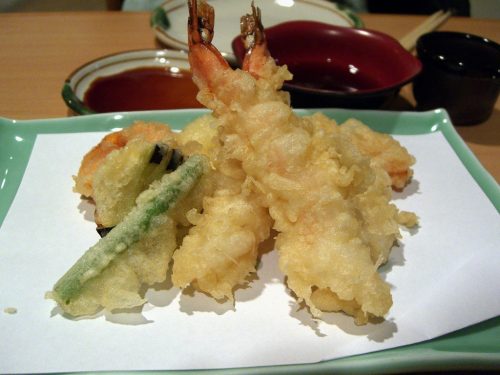 A plate of assorted tempura, including shrimp and vegetables, on a wooden table with dipping sauce in the background.