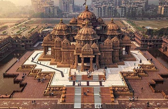 Aerial view of a large Hindu temple with multiple domes, intricate carvings, a courtyard, and modern buildings nearby.