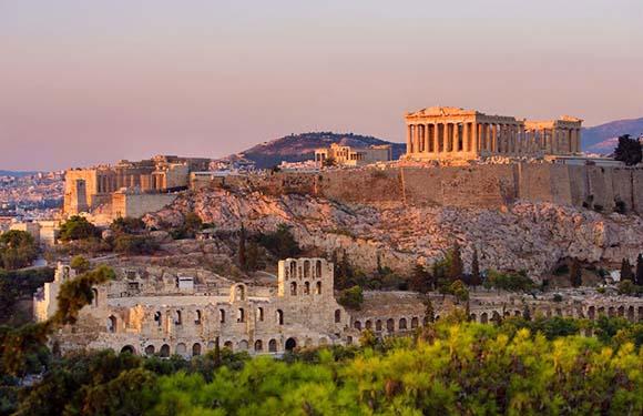 A scenic view of the Acropolis in Athens, Greece, during sunset with the Parthenon and other ancient ruins visible against a softly lit sky.