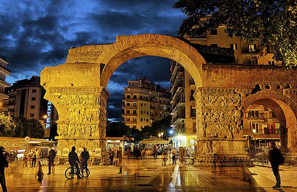 An image of an illuminated ancient Roman archway at night with people and modern buildings in the background.