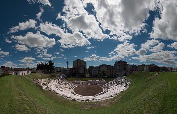 An ancient amphitheater with tiered seating and grassy slopes, under a cloudy sky, with modern buildings in the background.