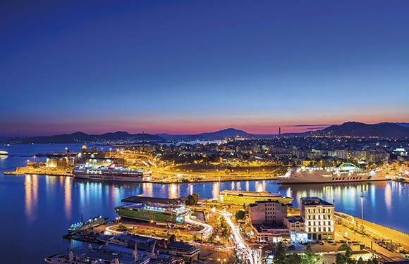 An image of a coastal cityscape at dusk with illuminated buildings and streets, and a calm sea reflecting city lights.