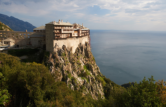 A building perched on a steep cliff overlooking a body of water with vegetation on the left side and a clear sky above.