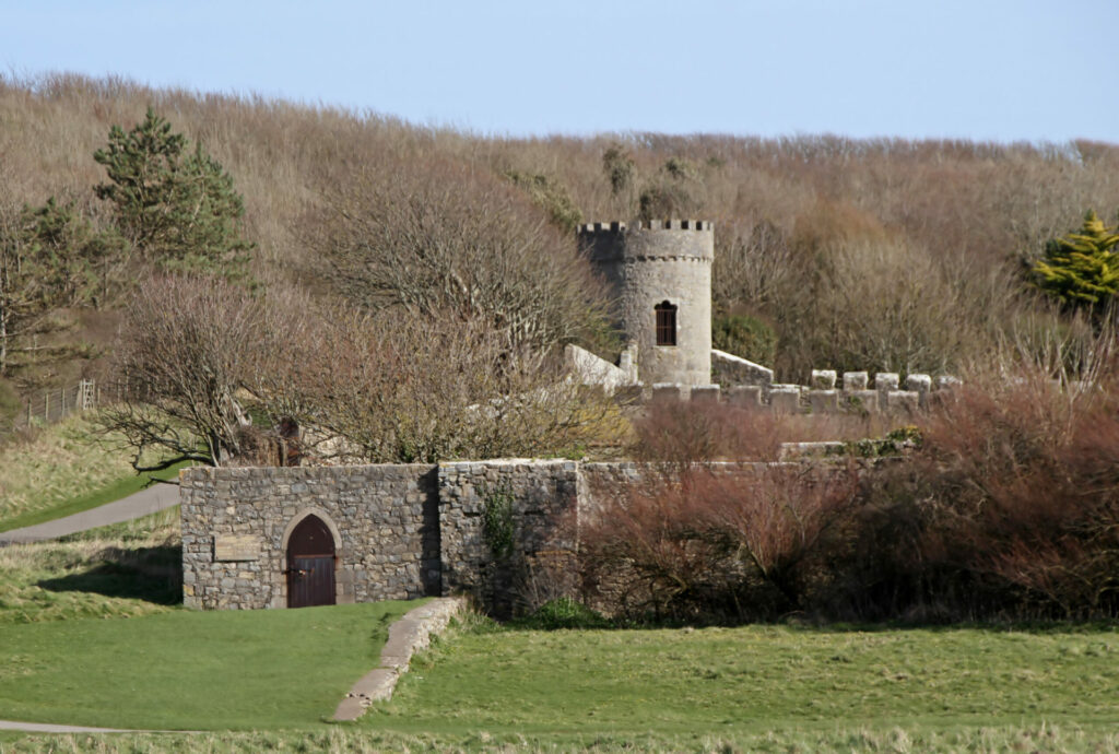 A small, stone castle with a turret surrounded by greenery under a clear sky.