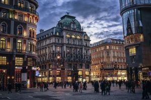 A bustling city square during twilight with people walking and historic buildings illuminated by street lights under a dramatic sky.