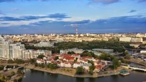 Aerial view of a cityscape with buildings of various heights, a river running through the area, and scattered greenery under a partly cloudy sky.