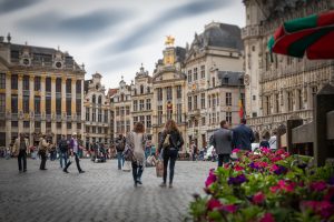 A bustling city square with people walking, historic buildings in the background, and vibrant flowers in the foreground.