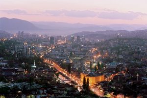 A cityscape during twilight with lights illuminating buildings and streets, and a hilly backdrop.