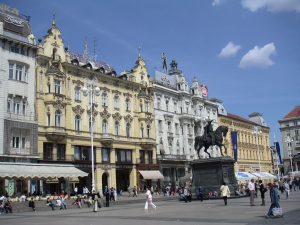 A city square featuring historical buildings, a statue of a mounted figure, and people walking under a clear, partly cloudy sky.