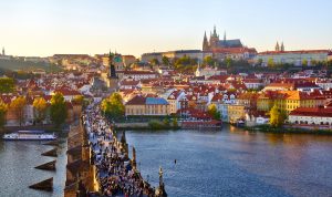 A panoramic view of a historic city at sunset with a large castle on a hill, a river in the foreground, and a bridge crowded with people.