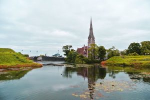 A scenic view of a church with a tall spire reflected in a calm river, surrounded by greenery and overcast skies.