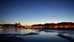 A panoramic view of a city skyline at dusk with lights reflecting on a body of water that has ice patches floating on the surface.