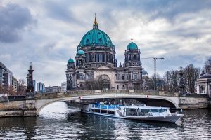 A photo shows the Berlin Cathedral, a bridge over the Spree River, a tour boat, and cloudy skies.