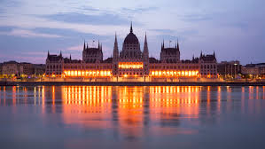 A nighttime view of the Hungarian Parliament Building with its reflection on the Danube River, lights illuminating the facade.