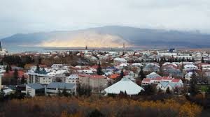 A city panorama featuring low-rise buildings, autumn trees, and a cloud-covered mountain.