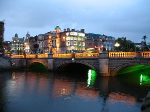 A nighttime cityscape with illuminated buildings and a bridge over a river, reflecting green lights on the water.